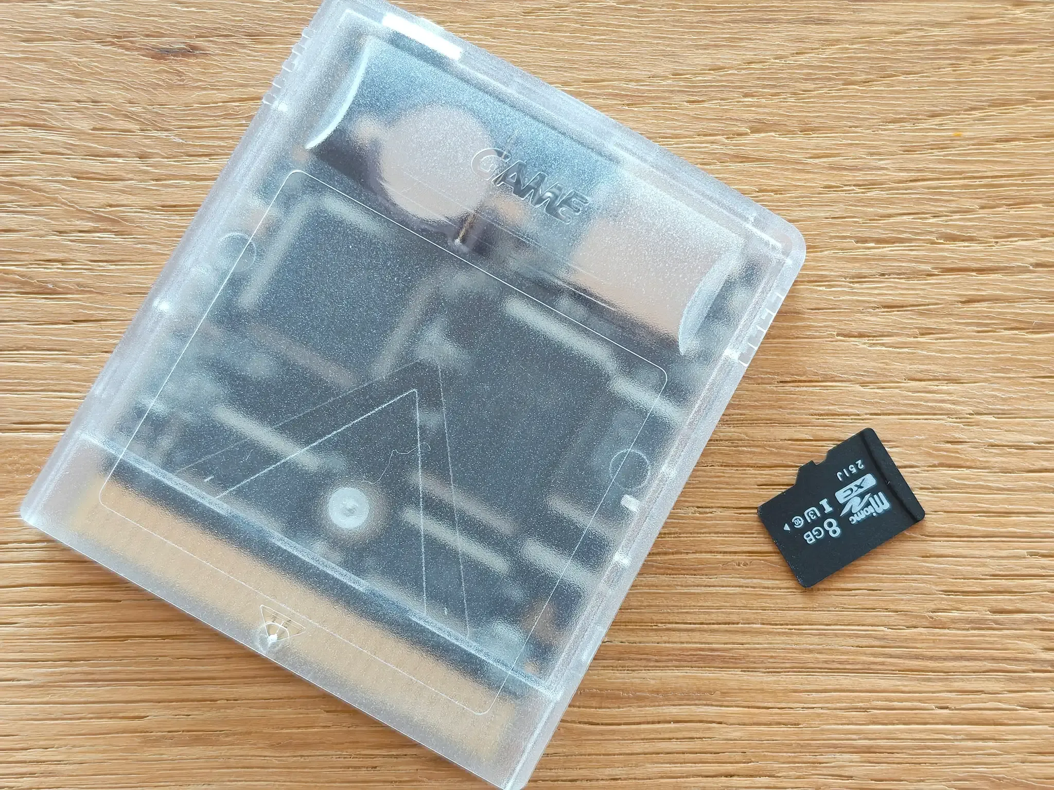 Transparente Game-Boy-Cartridge mit sichtbarer Platine neben einer schwarzen microSD-Karte auf Holztisch