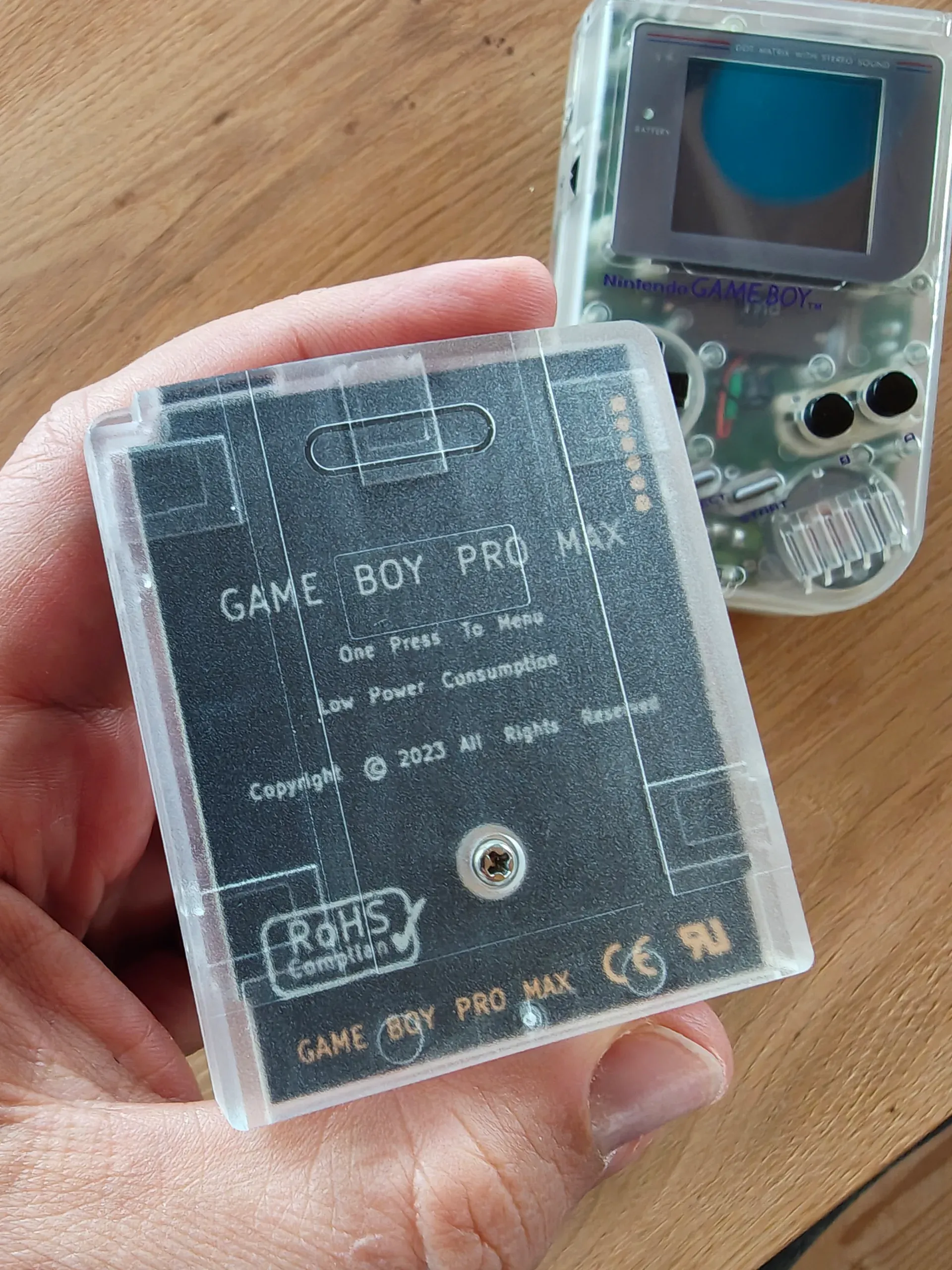 Hand hält transparente Game-Boy-Cartridge mit Aufdruck „GAME BOY PRO MAX“, dahinter ein durchsichtiger Game Boy auf Holztisch