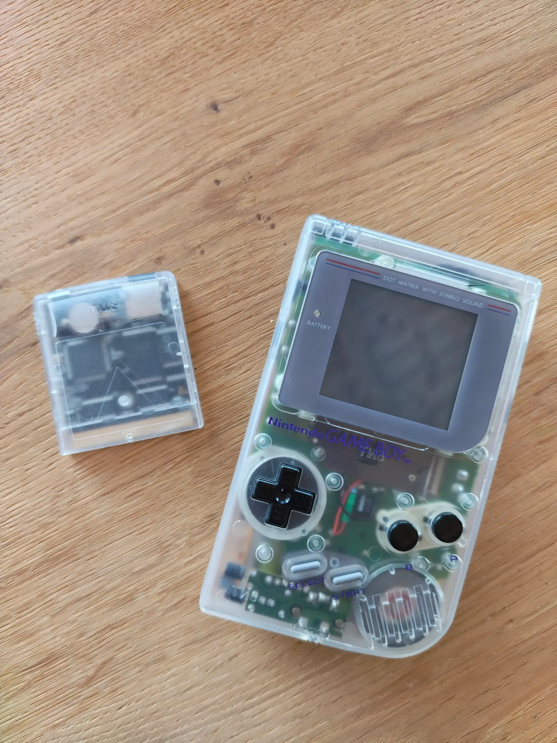 Transparenter Game Boy mit sichtbarer Platine neben einer durchsichtigen Spielmodul-Cartridge auf Holztisch