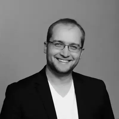 Lächelnder Mann mit Brille, Bart und dunklem Sakko über weißem Shirt in einem Schwarz-Weiß-Porträt