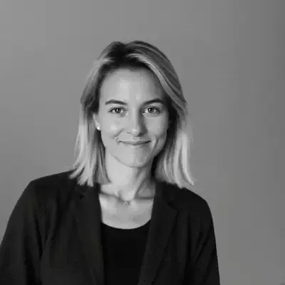 Schwarz-weisses Porträt einer lächelnden blonden Frau mit schulterlangem Haar und dunklem Blazer vor grauem Hintergrund